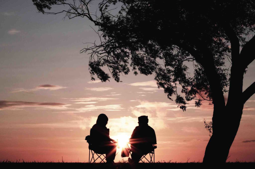 Two people sitting on camping chairs, next to a tree, all silhouetted by a dipping sun at dusk.