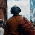 A shot from behind, of someone walking down a street - they are where headphone and carrying a backpack.